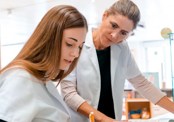 female blonde pharmacist helps and assists her young aprrentice trainee during studies of medical books A female blonde pharmacist helps and assists her young aprrentice trainee during studies of medical books